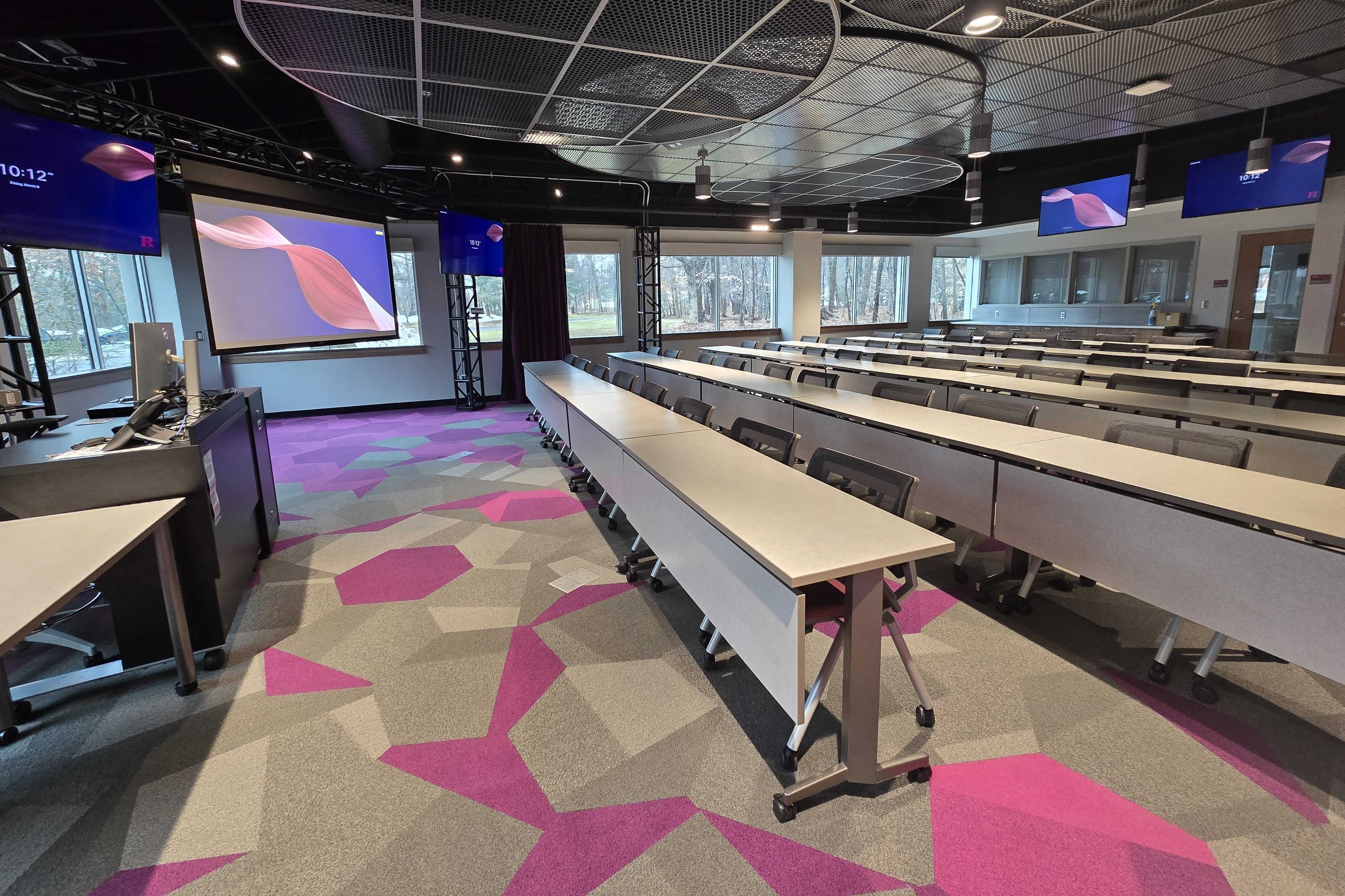 High technology classroom: rows of tables and chairs, projection screen in the front is down, side monitors and back monitors are on.