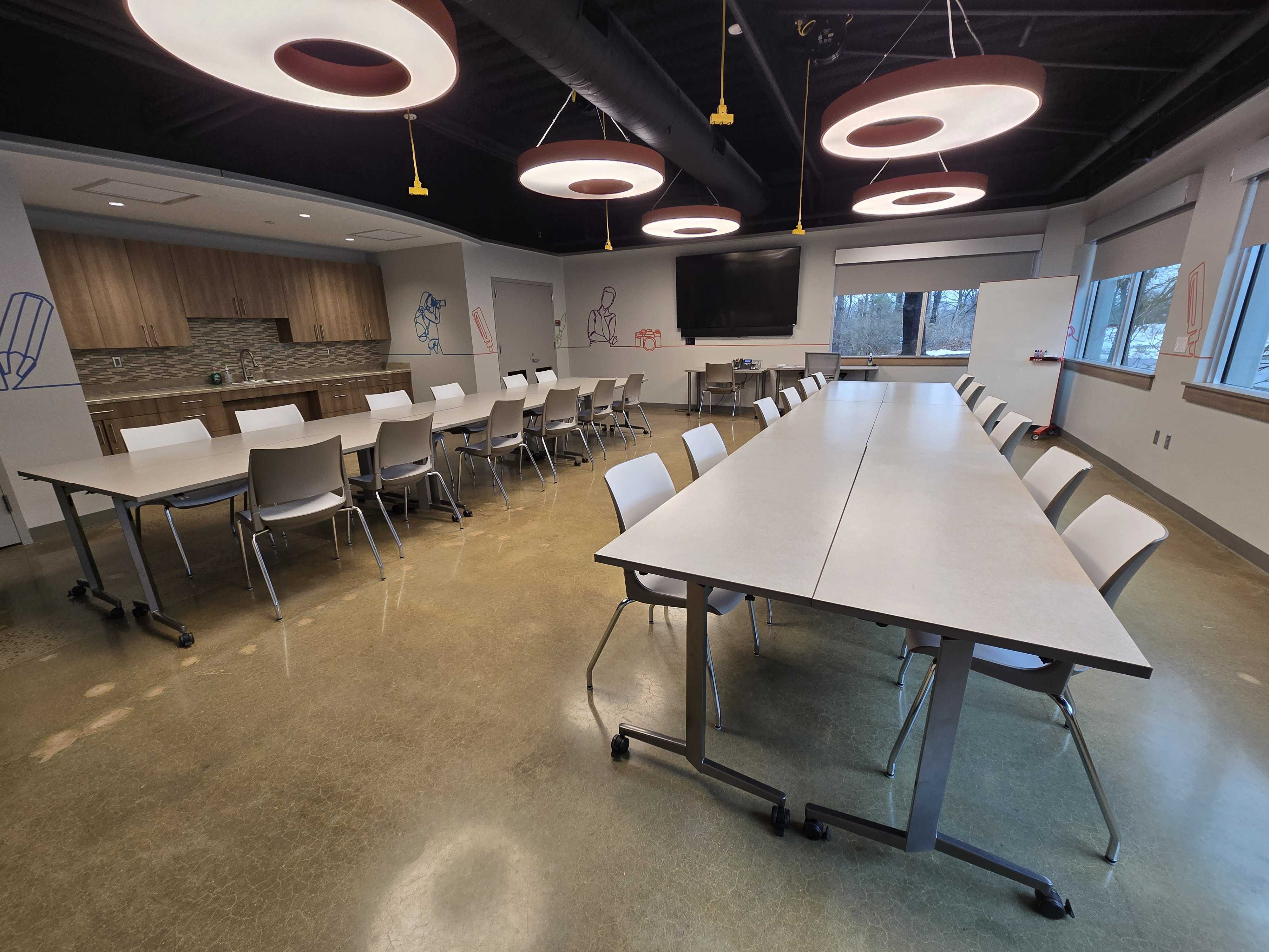 2 large groups of tables and chairs on either side of the room. Front TV monitor and a side sink area.