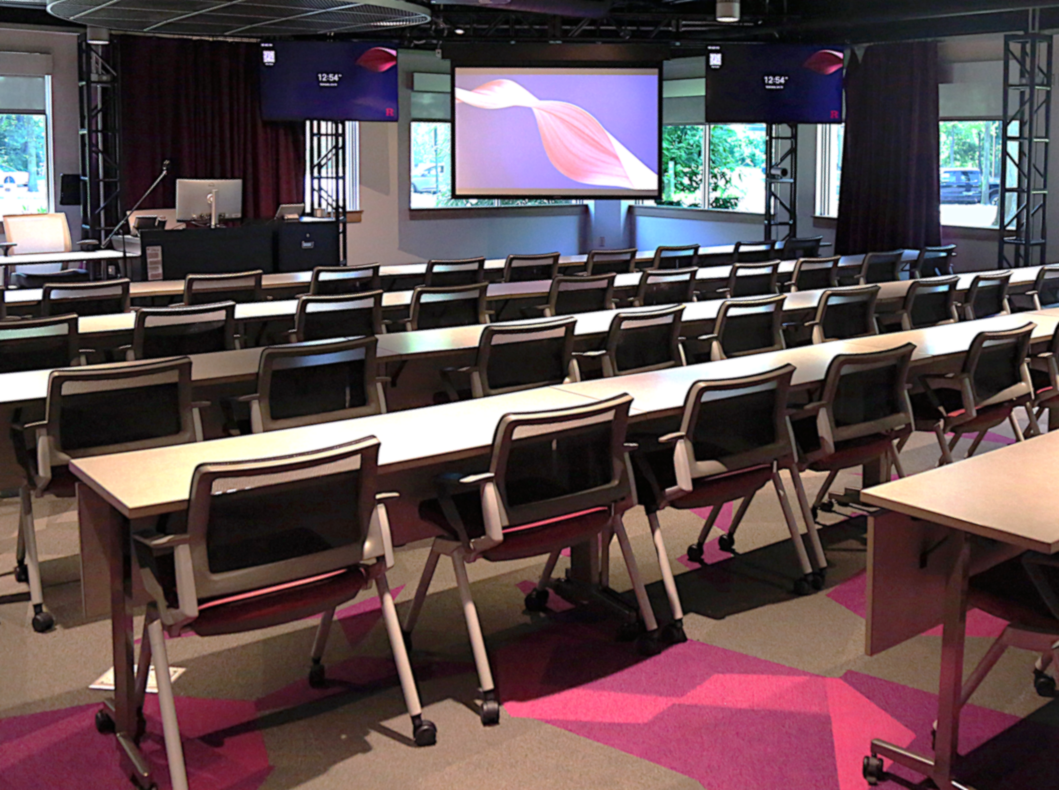 Classroom with rows of tables and chairs facing the front projection screen and side monitors